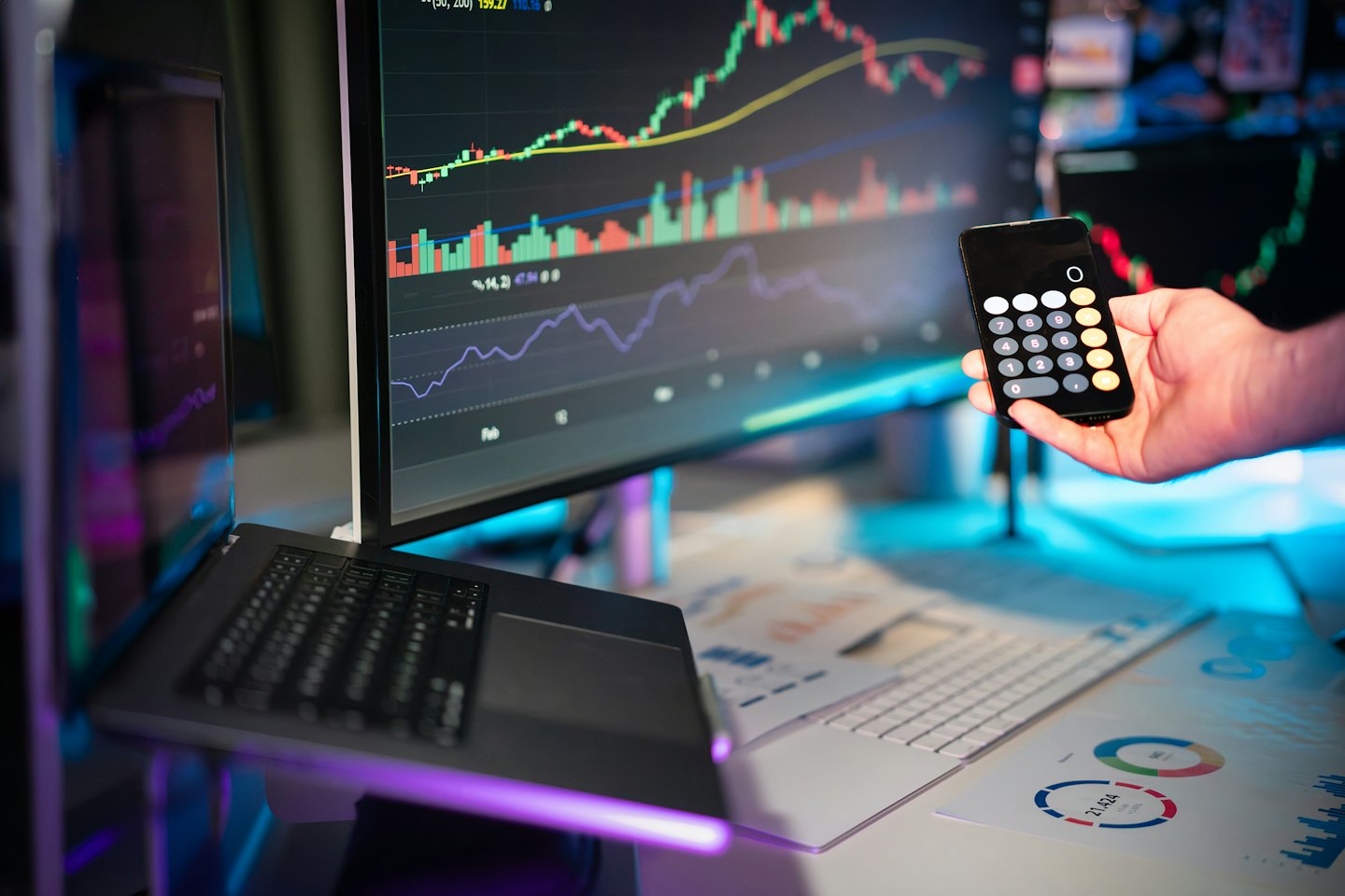 Hand håller telefon med kalkylator nära finansiell data på skärmar.